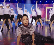 a woman in a black and silver dress is kneeling on the floor in front of a group of dancers wearing white hats