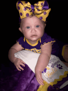 a baby wearing a purple and gold outfit holds a blanket