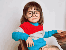 a little girl wearing glasses and a sweater sits on a chair