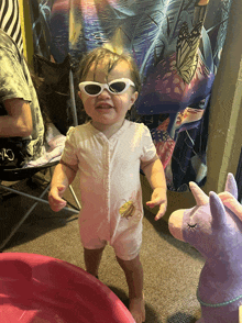 a baby wearing sunglasses stands next to a stuffed unicorn and a pink pool