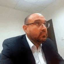 a man wearing glasses and a suit is sitting at a desk in a room .