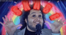 a man with a beard stands in front of a microphone with a rainbow in the background and the word skittles above his head