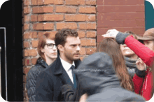 a man in a suit and tie is standing in front of a brick wall and a woman is taking a picture of him