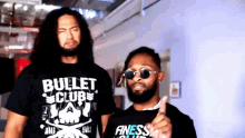 a man wearing a bullet club shirt is standing next to another man wearing sunglasses