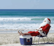 santa claus is sitting on a chair on the beach with his feet on a cooler .