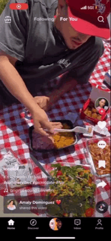 a man in a red hat sits on a checkered picnic blanket eating food