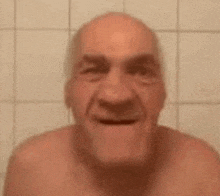 an elderly man without a shirt is making a funny face in front of a tiled wall .