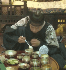 a man is sitting at a table with bowls of food and a fan and eating with a spoon and fork