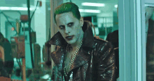 the joker is wearing a leather jacket and chains and is standing in front of a glass door .