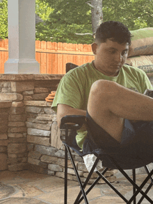 a man in a green shirt sits in a folding chair with his legs crossed and a bag of asphalt in the background