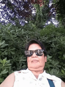 a woman wearing sunglasses takes a selfie in front of a bush