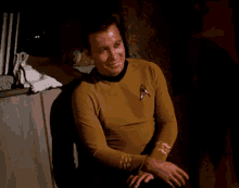 a man in a star trek uniform sits in a dark room smiling