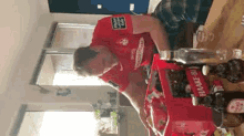 a man in a red shirt is sitting at a table with a box of hawker beer on it .