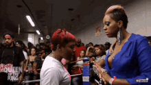 a woman in a blue shirt is talking to another woman in a boxing ring with the words queen of the ring on the wall