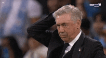 a man in a suit and tie scratches his head while watching a soccer game live