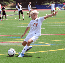 a boy in a white kia shirt kicking a soccer ball on a field