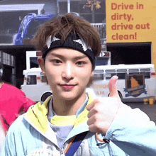 a young man wearing a headband gives a thumbs up in front of a sign that says drive in dirty drive out clean