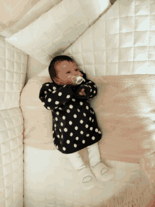 a baby in a black and white polka dot dress is laying in a crib with a pacifier in her mouth