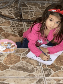 a little girl in a pink shirt is drawing on a piece of paper on the floor