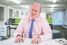 a man is sitting at a desk in an office wearing a pink shirt and blue tie .