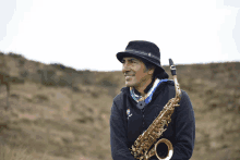 a man in a hat holds a saxophone in front of a hill