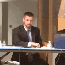 a man in a suit and tie is sitting at a table with a laptop and bottles of water .