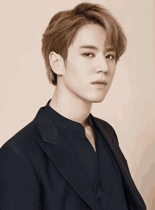 a young man wearing a black suit and a black shirt is standing in front of a beige background .