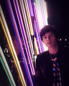 a young man wearing a t-shirt that says evil-s-de is standing in front of a wall of neon lights