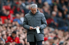 a man in a grey jacket is standing in front of a crowd holding a piece of paper in his hand .