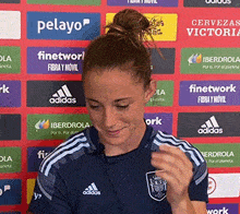 a woman in a blue adidas shirt stands in front of a wall of advertisements including pelayo and cervezas victor