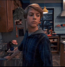 a young man in a blue plaid shirt stands in a kitchen