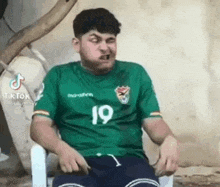 a man in a green shirt and shorts is sitting in a chair making a funny face .