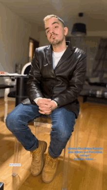 a man in a leather jacket sits on a clear chair with his hands folded