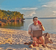 a man sitting on a beach with a laptop and a drink