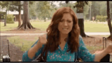 a woman with red hair and a blue shirt is sitting in a park with trees in the background .