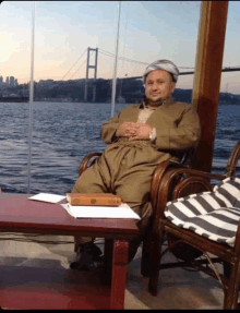 a man sits in a chair with a book on a table in front of him with a bridge in the background