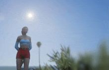 a woman in a blue top and red shorts stands in front of the sun