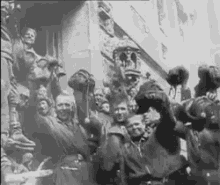 a black and white photo of a group of people standing in front of a building holding their hands up in the air .