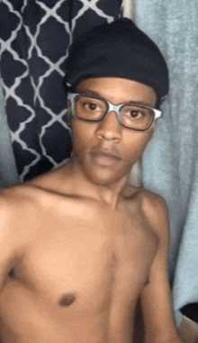 a shirtless man wearing glasses and a beanie is taking a selfie in front of a curtain .