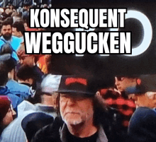 a man in a hat stands in front of a crowd with the words konsequent weggucken above him