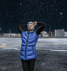 a man wearing sunglasses and a blue vest stands in a parking lot at night