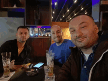 three men sitting at a table with glasses of beer and a cell phone