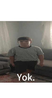 a young boy wearing a striped shirt and shorts is standing in front of a couch with the word yok on the bottom right