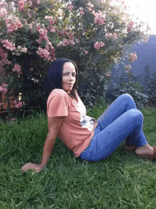 a woman is sitting in the grass in front of a bush of pink flowers