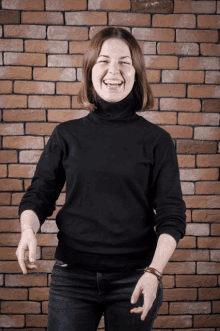 a woman wearing a black turtleneck sweater stands in front of a brick wall and smiles