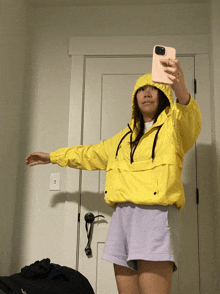 a woman wearing a yellow jacket and purple shorts takes a selfie with her phone