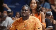 a man and woman are sitting in the stands at a basketball game . the man is wearing glasses and a nike shirt .