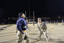 a man in a blue hoodie stands next to a man in a military uniform on a football field