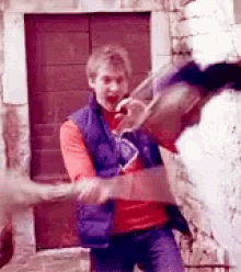 a man in a red shirt and a blue vest is standing in front of a brick wall holding a knife .