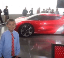 a boy in a blue shirt and red tie stands in front of a red sports car